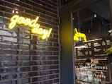 A neon sign with the words 'good day!' in bright yellow is mounted on a wall tiled with glossy black bricks. Beside it, a glass door reveals a coffee shop interior, with a hanging light fixture and a menu on the wall. The trading hours are listed on the door, and a person is seen working behind the counter.