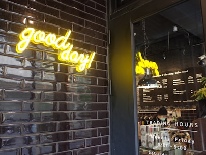 A neon sign with the words 'good day!' in bright yellow is mounted on a wall tiled with glossy black bricks. Beside it, a glass door reveals a coffee shop interior, with a hanging light fixture and a menu on the wall. The trading hours are listed on the door, and a person is seen working behind the counter.