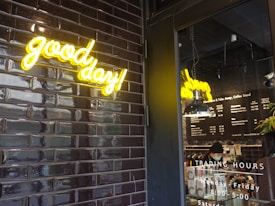 A neon sign with the words 'good day!' in bright yellow is mounted on a wall tiled with glossy black bricks. Beside it, a glass door reveals a coffee shop interior, with a hanging light fixture and a menu on the wall. The trading hours are listed on the door, and a person is seen working behind the counter.