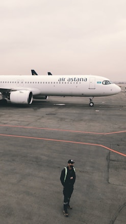 An airplane with the branding 'air astana' is parked on a tarmac. The aircraft is white with a few blue and gold accents, and the scene includes a cloudy sky. A person wearing a black outfit and a cap stands on the tarmac in front of the plane, with red lines marked on the ground.