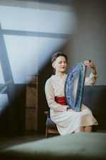 A person sits on a chair indoors, holding a large, colorful handheld drum-like instrument with one hand. The setting is softly lit, with muted colors and a large window in the background, casting natural light across the scene. The person has a serene expression, wearing a light-colored dress with a bright red belt.