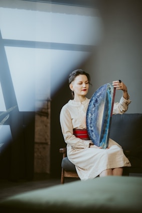 A person sits on a chair indoors, holding a large, colorful handheld drum-like instrument with one hand. The setting is softly lit, with muted colors and a large window in the background, casting natural light across the scene. The person has a serene expression, wearing a light-colored dress with a bright red belt.