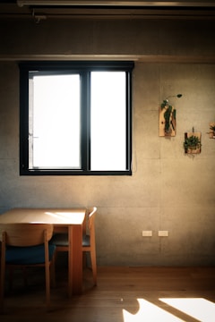 A minimalist interior with a concrete wall and a large window allowing bright natural light to stream in. Two wooden chairs are neatly placed around a wooden table. There are two small wall-mounted plant decorations on the wall to the right of the window.