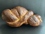 A single loaf of braided bread with a golden-brown crust placed against a dark background. The bread has a shiny surface, indicating it has been glazed.