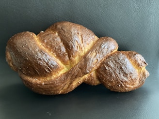 A single loaf of braided bread with a golden-brown crust placed against a dark background. The bread has a shiny surface, indicating it has been glazed.