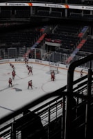 An ice hockey match is taking place in an arena with empty spectator seats in the background. Several players in red uniforms are on the ice, engaged in the game. The arena features advertisements and bright lighting strips along the staircases. The atmosphere is tense and competitive.