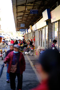 A bustling street market with various people walking and cycling. Stalls displaying goods are lined up along the right side under a large roof. Several signs hang from the ceiling, indicating services like a toilet and parking. Many shoppers engage with vendors, and some are wearing masks.