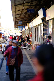 A bustling street market with various people walking and cycling. Stalls displaying goods are lined up along the right side under a large roof. Several signs hang from the ceiling, indicating services like a toilet and parking. Many shoppers engage with vendors, and some are wearing masks.