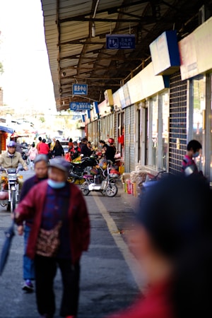 A bustling street market with various people walking and cycling. Stalls displaying goods are lined up along the right side under a large roof. Several signs hang from the ceiling, indicating services like a toilet and parking. Many shoppers engage with vendors, and some are wearing masks.