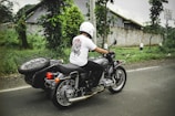 A person wearing a helmet and a white t-shirt is riding a motorcycle with a sidecar along a road. The sidecar has a spare tire mounted on it. The surroundings include green foliage and a concrete wall in the background. The weather appears overcast.