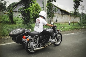 A person wearing a helmet and a white t-shirt is riding a motorcycle with a sidecar along a road. The sidecar has a spare tire mounted on it. The surroundings include green foliage and a concrete wall in the background. The weather appears overcast.