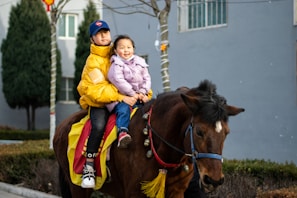 Two children are sitting on a brown horse. The older child is wearing a yellow jacket and a blue cap, while the younger child is in a purple jacket. The horse is adorned with a colorful red and yellow blanket, and decorations hanging from its bridle. In the background, there are trees and a building with a window.