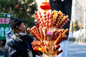 A street vendor is standing beside an assortment of traditional candied fruits on skewers, displayed in a colorful and vibrant arrangement. The vendor is wearing a black coat and hat, with a medical mask hanging beneath the chin. The background features some blurred elements, including a green sign with writing and a large red lantern, suggesting a busy area or market.
