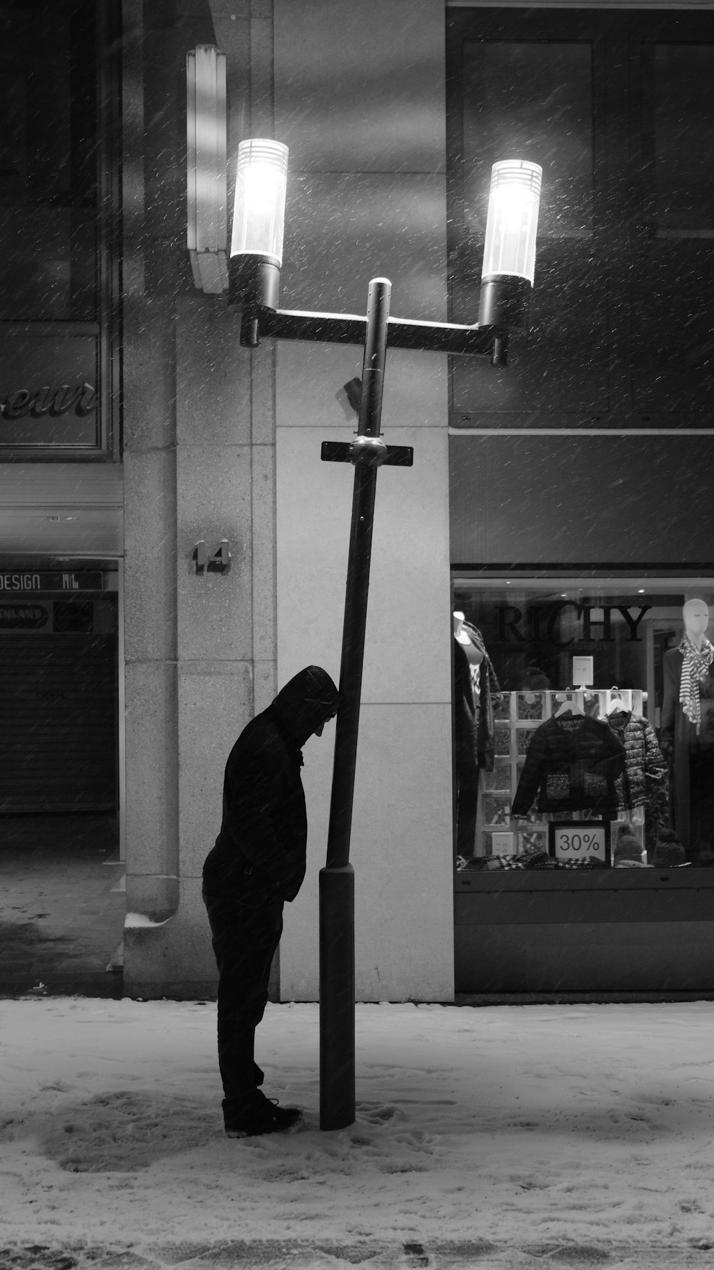 grayscale photo of man in black hoodie standing near glass window