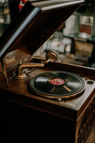 A vintage gramophone with an open wooden lid, featuring a brass tonearm and a black vinyl record with a red label. The background is softly blurred, hinting at an antique or rustic setting.