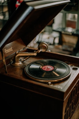 A vintage gramophone with an open wooden lid, featuring a brass tonearm and a black vinyl record with a red label. The background is softly blurred, hinting at an antique or rustic setting.