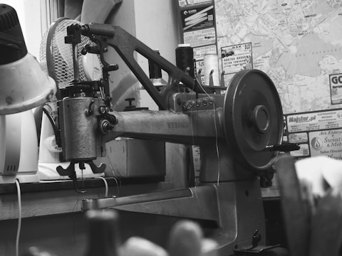 A vintage sewing machine on a workbench surrounded by various tools and materials. A fan and several spools of thread are visible in the background alongside a wall covered with maps and advertisements. The atmosphere conveys a sense of timeworn functionality and industriousness.