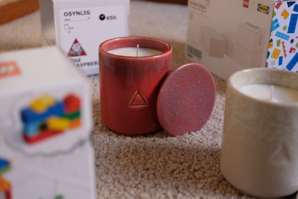 A red ceramic candle with a geometric pattern sits on a carpeted floor, partially covered by its matching lid. Another white candle in a textured jar is nearby, along with colorful packaging boxes labeled IKEA that are slightly blurred in the background.