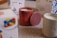 A red ceramic candle with a geometric pattern sits on a carpeted floor, partially covered by its matching lid. Another white candle in a textured jar is nearby, along with colorful packaging boxes labeled IKEA that are slightly blurred in the background.