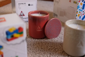 A red ceramic candle with a geometric pattern sits on a carpeted floor, partially covered by its matching lid. Another white candle in a textured jar is nearby, along with colorful packaging boxes labeled IKEA that are slightly blurred in the background.