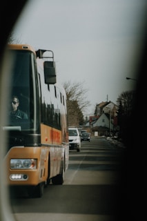 A large orange bus is driving on a suburban street alongside other vehicles, including a white car. The road is lined with houses and trees, and the atmosphere seems calm and routine.