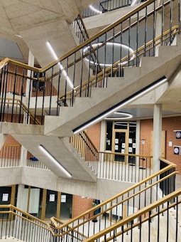 A modern, multi-level staircase with geometric design elements, featuring concrete steps and railings with wooden accents. Bright, linear LED lights illuminate the structure, creating an industrial yet elegant look. Brick walls and glass doors are visible in the background.