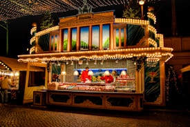 A brightly lit and decorative street food stall at night, resembling a charming, festive house with numerous lights outlining its structure. Inside, there are two people dressed in red selling various treats and snacks. The background features an elaborate painting of hills and trees, and the stall has a painted facade with illuminated signage. Other stalls and string lights can be seen nearby as well.