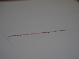 A minimalistic design featuring a piece of paper with the text 'Typography is what language looks like.' written in red ink. The focus on the text gives a stylish and modern feel with ample white space around it.