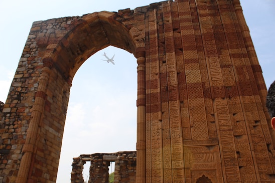 An ancient stone archway with intricate carvings and patterns stands tall against a bright sky. Through the arch, a commercial airplane is visible, suggesting a juxtaposition of ancient and modern elements. The stones show varied earth tones, with detailed inscriptions and a partially ruined structure in the background.