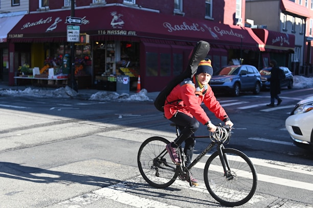A person rides a bicycle through an urban intersection, wearing a bright red jacket and a colorful beanie. They carry a large case on their back, suggesting it might hold a musical instrument. Behind them is a store with a red awning labeled 'Rachel's Garden', displaying various fruits and vegetables.