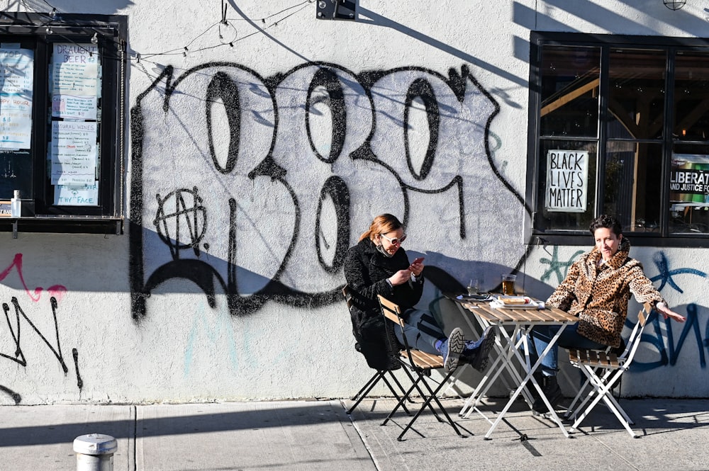 man in black jacket sitting on folding chair beside wall with graffiti during daytime