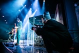 A stage setup with musicians and a videographer. The musicians include a guitarist and a keyboardist, captured mid-performance. A person wearing a headset operates a camera from the side, focusing on the musical performance. Bright stage lights and a darkened background create a vibrant and dynamic environment.