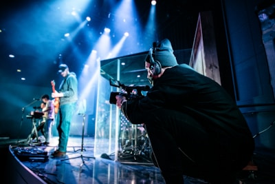 A stage setup with musicians and a videographer. The musicians include a guitarist and a keyboardist, captured mid-performance. A person wearing a headset operates a camera from the side, focusing on the musical performance. Bright stage lights and a darkened background create a vibrant and dynamic environment.