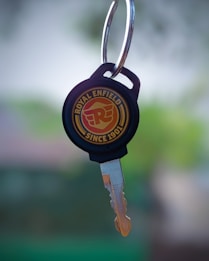 A close-up of a metallic key with a round black plastic grip, displaying the Royal Enfield logo and the text 'Since 1901'. The background is blurred, showcasing a mix of green and neutral tones.