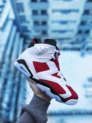 A person wearing a grey sweatshirt holds up a red, white, and black sneaker against a blurred backdrop of a building, likely taken from a height. The shoe features intricate leather designs with perforations for breathability.