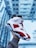 A person wearing a grey sweatshirt holds up a red, white, and black sneaker against a blurred backdrop of a building, likely taken from a height. The shoe features intricate leather designs with perforations for breathability.