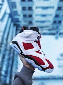 A person wearing a grey sweatshirt holds up a red, white, and black sneaker against a blurred backdrop of a building, likely taken from a height. The shoe features intricate leather designs with perforations for breathability.