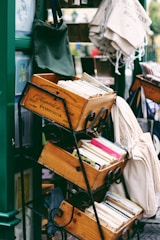 Several stacked wooden crates are filled with books, including some visible colorful spines. The scene is part of an outdoor bookshelf display with fabric bags hung on an adjacent stand.