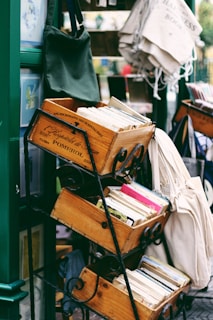 Several stacked wooden crates are filled with books, including some visible colorful spines. The scene is part of an outdoor bookshelf display with fabric bags hung on an adjacent stand.