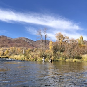 Two people are fly fishing in a river surrounded by autumn foliage. The background features hills with trees displaying a mixture of yellow, brown, and green leaves under a clear blue sky with wispy clouds.