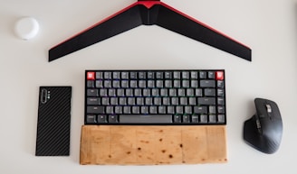 A mechanical keyboard with a wooden wrist rest is centered on a white surface. Above it is a red and black geometric object. On the left, there is a smartphone with a carbon fiber case, and on the right, a black ergonomic wireless mouse. A small white round item is also seen in the top left corner.