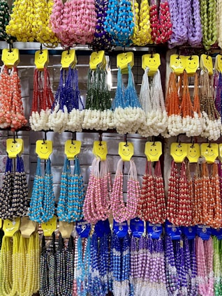 A colorful display of beaded necklaces is hanging on a wall. The necklaces are organized by color and type, including red, blue, green, yellow, purple, orange, pink, and white variations. Each set is hung on hangers adorned with smiley faces, creating a cheerful and inviting presentation.