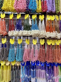 A colorful display of beaded necklaces is hanging on a wall. The necklaces are organized by color and type, including red, blue, green, yellow, purple, orange, pink, and white variations. Each set is hung on hangers adorned with smiley faces, creating a cheerful and inviting presentation.