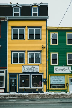A vibrant yellow and green row of townhouses featuring quaint storefronts. The building on the left is yellow with detailed navy blue trimming and a sign reading 'Posie Row'. To the right, a green building with white-framed windows displays a sign for 'Elaine's Books.' Snow is piled on the sidewalk below.