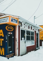 A small pizza shop with a weathered exterior, featuring a prominently placed ATM machine. The storefront has a sign reading 'Sal's Pizza' and is situated amidst a snowy landscape. There are various signs and stickers on the door and windows.