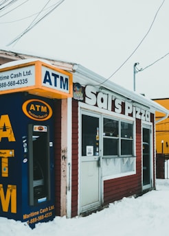 A small pizza shop with a weathered exterior, featuring a prominently placed ATM machine. The storefront has a sign reading 'Sal's Pizza' and is situated amidst a snowy landscape. There are various signs and stickers on the door and windows.