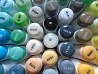 A collection of various colored marker caps arranged in a circular formation, displaying a range of color codes. The caps are tightly packed together, showcasing a spectrum of shades from blues and greens to grays and browns.