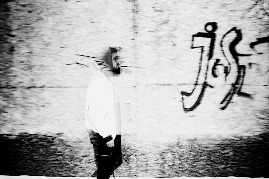 A black and white street photograph showing a lone figure walking past a graffiti-covered wall, with strong contrast and deep shadows.