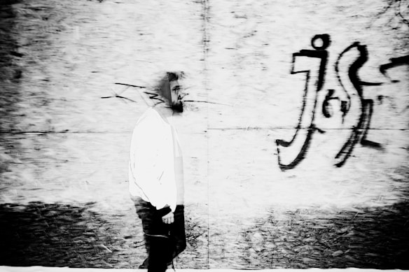 A black and white photograph depicting a person walking along a wall with graffiti. The image has motion blur, particularly around the person's face, giving it a dynamic and slightly surreal appearance. The graffiti adds an urban and edgy element to the scene.