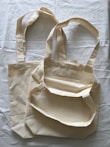 Two beige cloth bags with handles laid flat on a slightly crumpled fabric surface. The bags have a simple, unadorned design and appear to be made of cotton or canvas material.